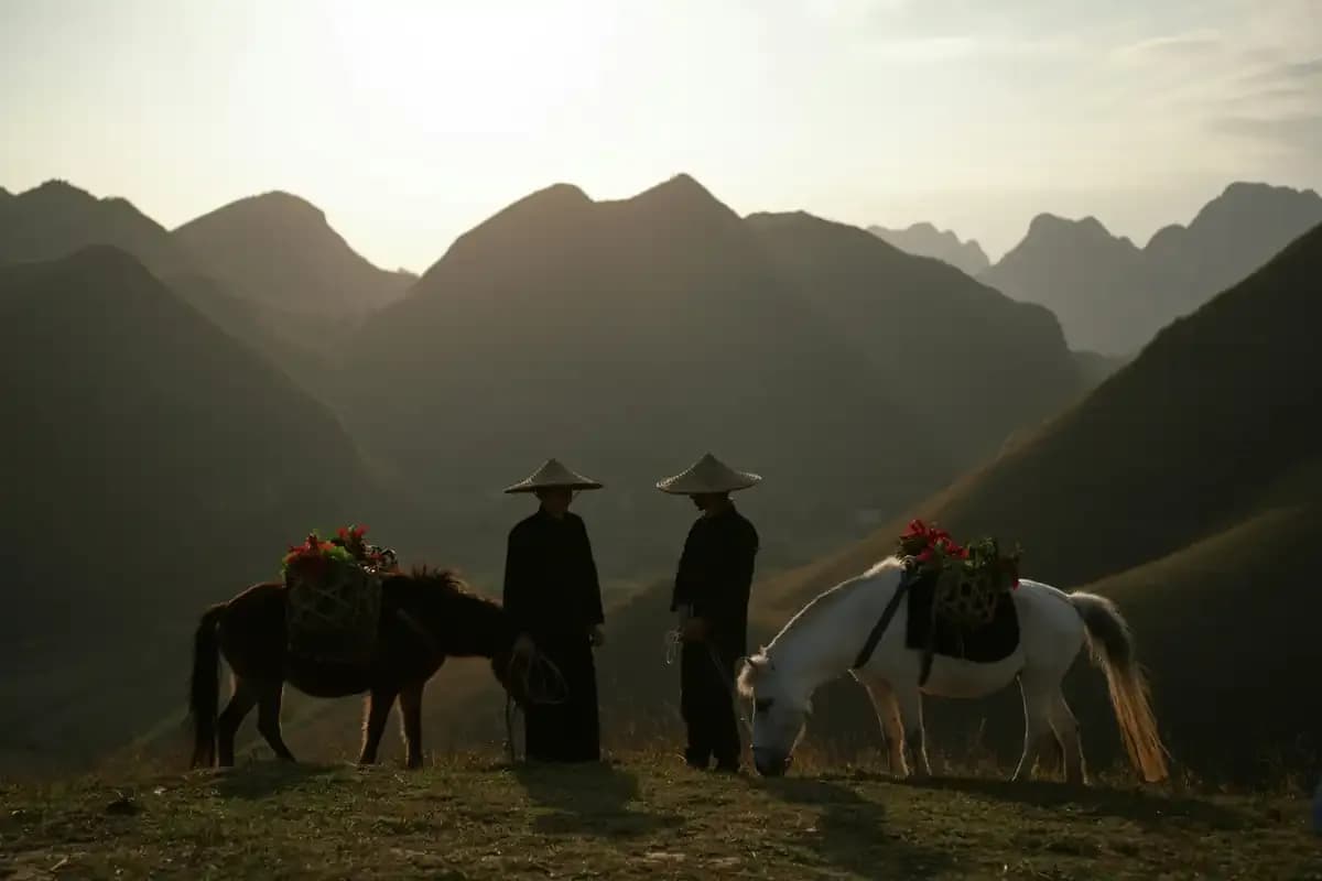 Two people standing with laden horses on a ridge before layered mountains at dusk