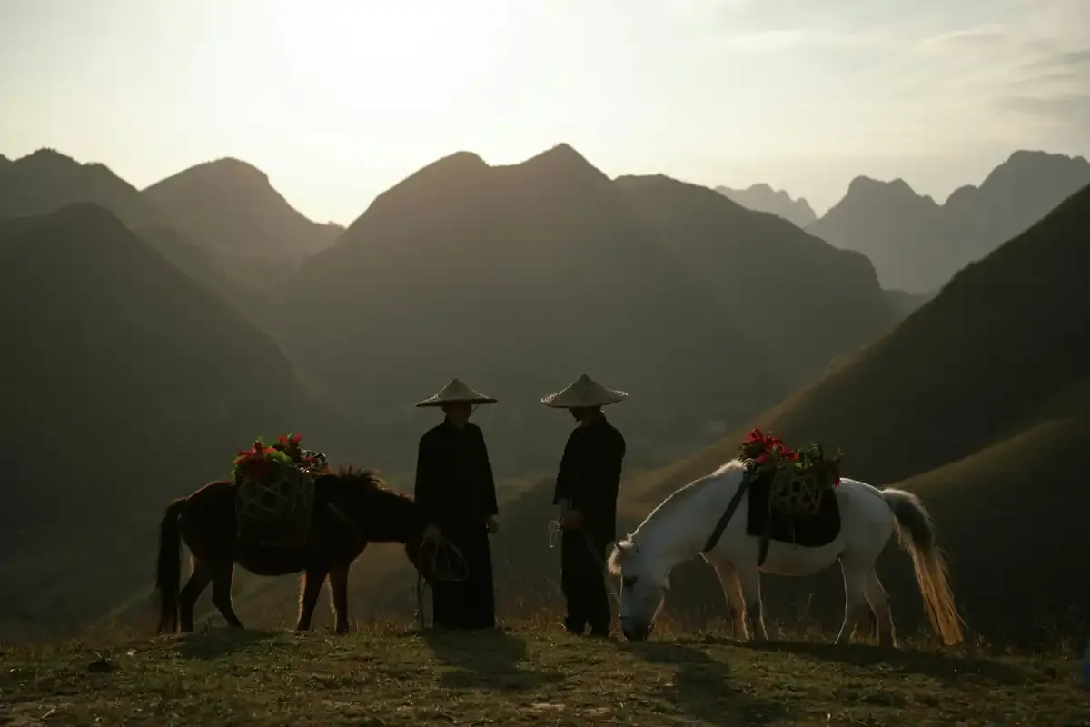 Two people standing with laden horses on a ridge before layered mountains at dusk